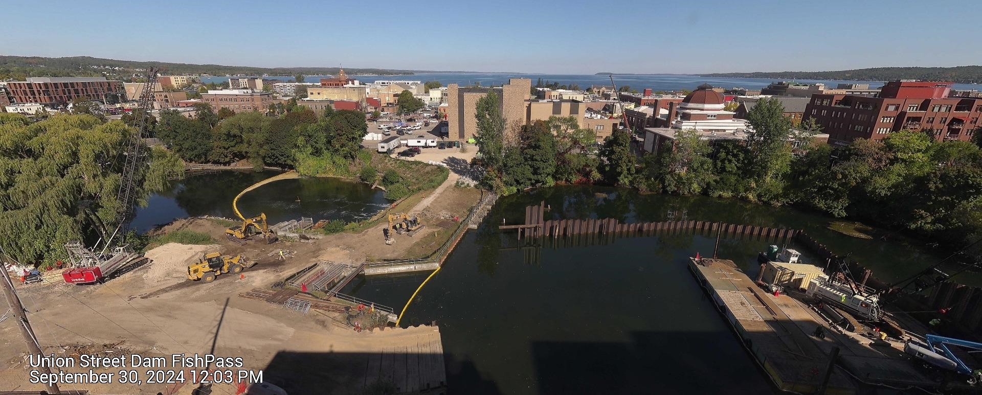 Aerial view of the Union Street Dam FishPass construction site, taken on September 30, 2024, at 12:03 PM. The image shows construction equipment, including excavators and cranes, on-site near the river. A yellow containment boom is deployed in the water, and part of the river is sectioned off with sheet piling. In the background, the city skyline with various buildings and a view of the bay can be seen under a clear blue sky.