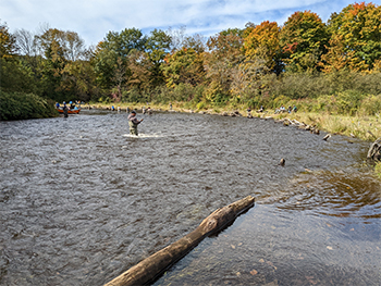 River curving up and to the left.  A person is fishing in the middle and several people in a canoe are rounding the bend at the back-left